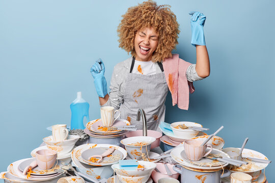 Positive housewife dances near sink full of dirty dishes with leftover food wears striped apron and rubber gloves does housework isolated over blue background. Daily domestic chores concept.