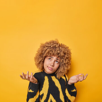 Unaware hesitant woman with curly hair spreads palms feels confused cannot decide on something wears casual jumper doesnt know what to do wears casual jumper isolated over yellow background.