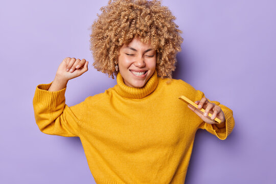 Waist up shot of carefree young woman dances with mobile phone dressed in warm yellow jumper has joyful expression has eyes closed isolated over purple background moves with rhythm of music.