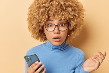 Portrait of curly haired woman stares through eyeglasses holds mobile phone reacts to shocking news sends text messages wears blue turtleneck isolated over beige background. Human reactions.