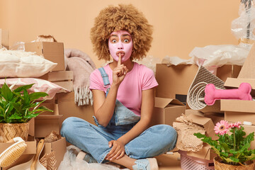 Surprised curly haired female apartment owner applies beauty facial mask sits crossed legs surrounded by cardboard boxes and personal belongings isolated over beige background. Relocation concept