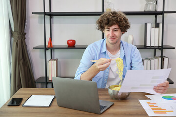 Young caucasian currly hair man sitting on table and eating tasty and instant noodle for lunch in the working room with chopsticks and computer. Unhealthy and cheap food lifestyle conceptncept.