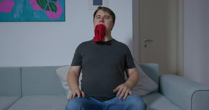Strange young man with a red nose is sitting on the couch and chewing a sock, he has a vomiting urge, then leaves away, front view. Person suffers from sleepwalking.