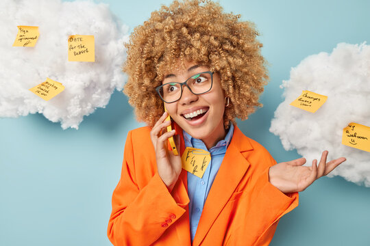 Positive businesswoman makes phone call shrugs shoulders and smiles broadly keeps smartphone near ear dressed in formal ornage jacket poses against blue background with memo sticky notes around