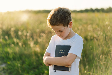 Christian boy holds bible in her hands. Reading the Holy Bible in a field during beautiful sunset....