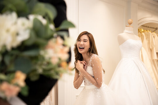 Asian woman in a bridal gown bright smiling. The groom wearing black suit holds a white bouquet of flowers to surprise the bride who stand in the fitting room. Concept wedding best wishes good day.
