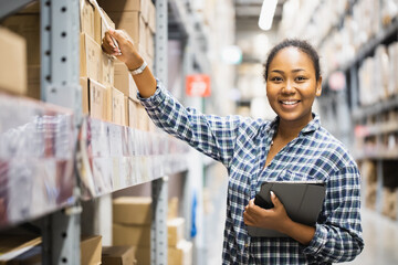 Portrait of young attractive African American woman auditor or trainee staff work looking up stocktaking inventory in warehouse store by computer tablet and stylus near products shelf. Audit concept
