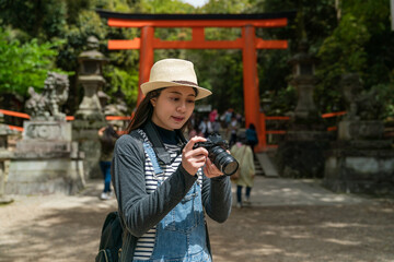 Asian Chinese woman photographer looking at her slr camera and checking photos near red torii gate...
