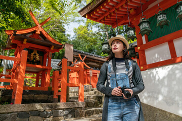 attractive asian Chinese girl photographer looking into distance at historic buildings while visiting Kasuga taisha near a red shinto shrine miniature in nara japan on sunny day