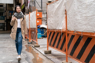 full length of Asian Japanese girl pedestrian passing by a construction site with orange safety barrier fence while walking on street in osaka city japan