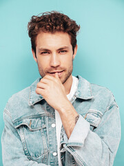 Portrait of handsome confident stylish hipster lambersexual model with curly hairstyle. Sexy man dressed in jeans jacket. Fashion male isolated on blue wall in studio