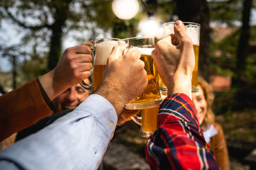 Cheerful friends celebrating toasting with beer mugs sitting outdoor - cropped shot - closeup on the hands