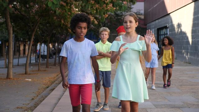 Cute preteen girl talking friendly with her african american friend while walking together along city street on summer day. High quality 4k footage
