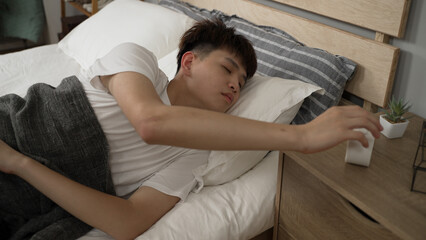 asian young man hitting the snooze button on the alarm clock and keep sleeping on bed in the morning in the bedroom at home.