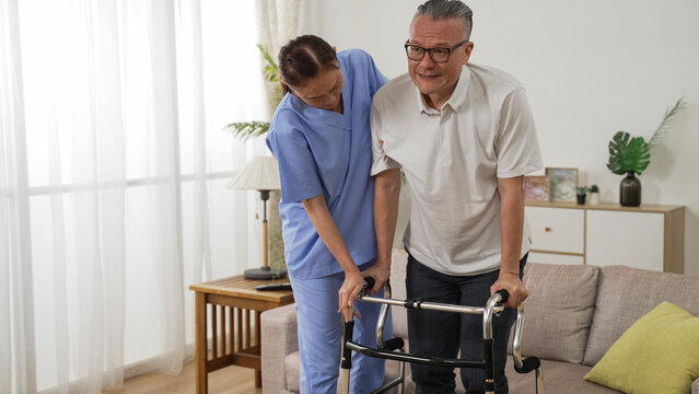 Asian Senior Stroke Patient Undergoing Rehab Exercise With A Walker At Home. The Woman Nursing Aide Assists Him During Home Visit