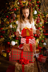 A cute child prepared a bunch of red gift boxes with ribbons for his friends and family stands against the backdrop of a beautiful Christmas tree