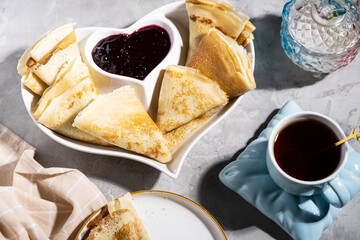thin pancakes with jam in a heart-shaped plate with tea on a gray background