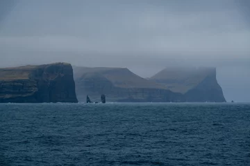 Fotobehang Impressive steep cliffs and rocky coastline silhouette coast of Faroe Islands in Atlantic Ocean on stormy grey day with clouds, lighthouse and hills seen from cruiseship cruise ship liner with spray © Tamme