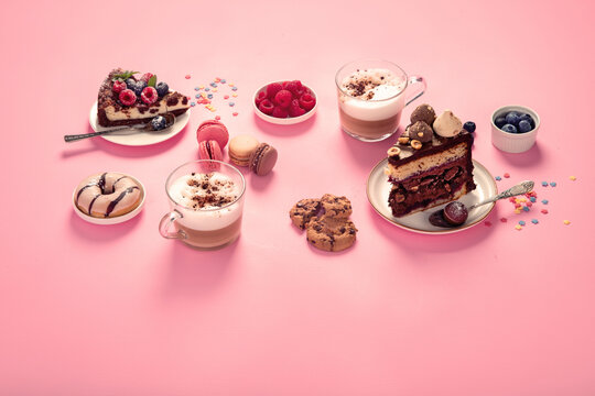 Table with various cookies, donuts, cakes, coffee cups on pink background.