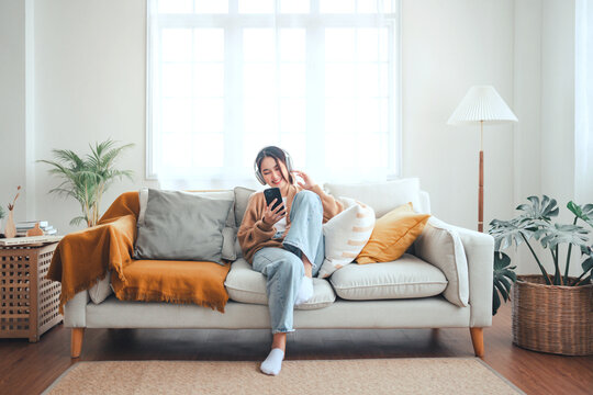 Happy asian woman listening to music from mobile phone while sitting on the the sofa at homes, Smiling girl relaxing with headphones in morning, Time to relax. copy space.