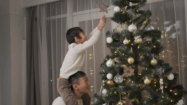 Happy farther lifting little son to put top star on christmas tree at home