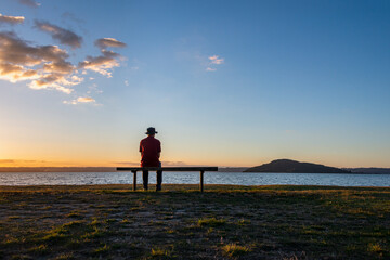 Fototapeta premium Man sitting alone by lake Rotorua at sunset, Mokoia Island in the distance, Rotorua.