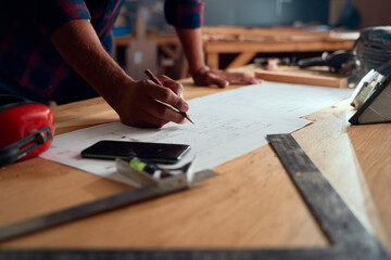 Close-up of mid adult man writing on paper next to mobile phone and tools in woodworking factory