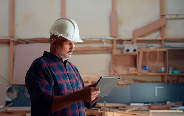 Focused mid adult man in safety helmet using digital tablet in woodworking factory