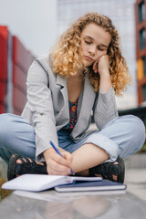 Pensive woman with curly hair taking notes, writing resume on notepad sitting on the street.