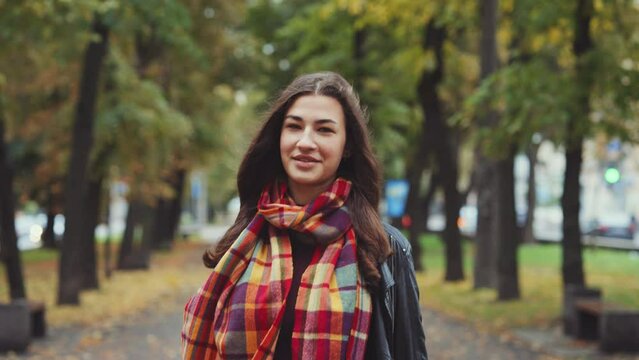 Young woman wearing leather jacket with checkered scarf walking in autumn city, looking at camera, blurred trees background. Tracking shot of happy person outside. Concept of joy