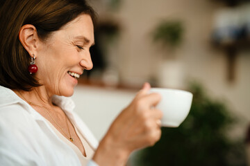 White mature woman in shirt smiling and drinking coffee in cafe indoors