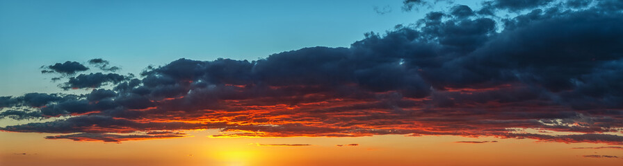 Setting sun painting red the bottom of a large gray cloud. Dramatic sky at golden hour. Bright vivid sunset sky panoramic shot. Picturesque wide skyscape at evening. Weather forecast.