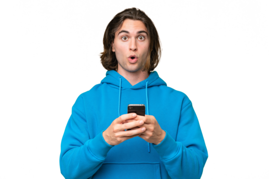 Young handsome man isolated on green chroma background looking at the camera while using the mobile with surprised expression