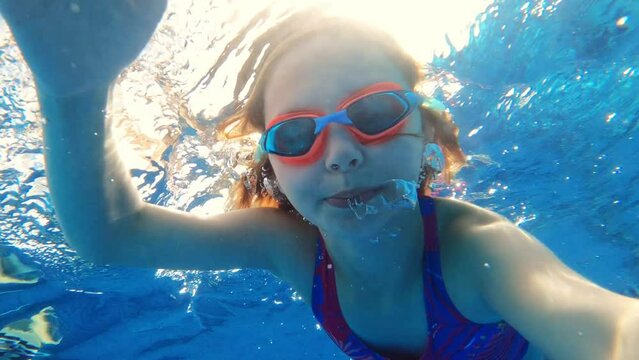 Cute little girl with goggles diving and swimming underwater in a pool. Kid taking selfie in water.