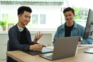 Two happy creative man looking at laptop screen and discussing online project at graphic studio