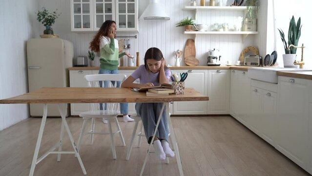 Unhappy Caucasian girl doing extracurricular work sits at table listening as mother swears talking on phone. Nervous woman calling tutor and screaming because of daughter's poor performance at school
