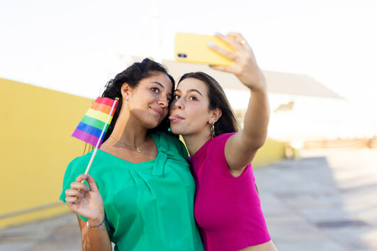 A beautiful lesbian young couple embraces and holds a rainbow flag. Girls taking selfie photo