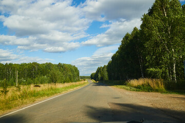 Fototapeta premium Landscape with road going into perspective, forest with trees on the roadsides, yellow fields and blue sky with cumulus white clouds