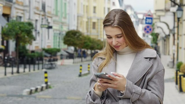 Smiling oung woman wearing casual gray coat walking writing sms online with smartphone. Dreamy stylish woman chatting at street, blurred old buildings on background. Mobile addiction concept
