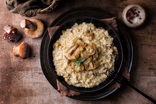 Risotto with porcini and champignon mushrooms in a black plate on brown background, directly above.