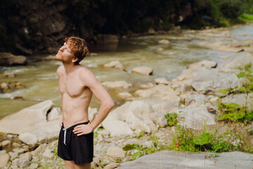 Fototapeta premium White young man wearing swimming trunks doing workout while resting by river