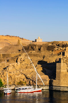 Egypt, Aswan Governorate, Aswan, Sailboats floating on bank of Nile river with Mausoleum of Aga Khan in background