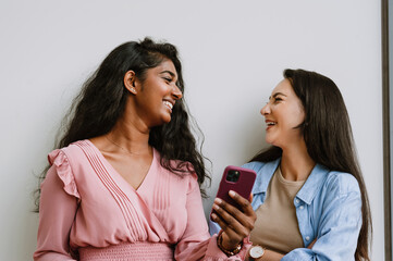 Two multinational women using cellphone while standing by wall outdoors