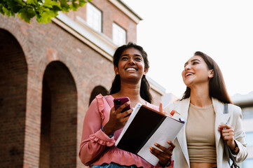 Multinational women smiling and using cellphone while walking outdoors