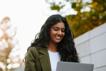 Brunette indian woman smiling while working with laptop on city street