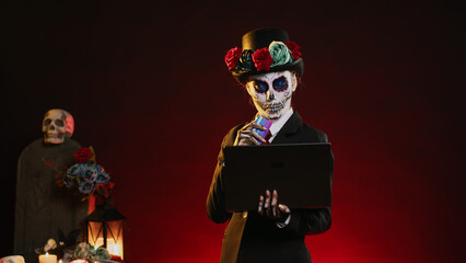 La cavalera catrina doing online shopping on laptop, using credit card to pay electronic transaction. Santa muerte making purchase on internet with pc, wearing death costume. Handheld shot.