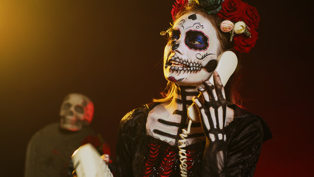 Goddess of death holding landline phone in studio, talking on office telephone with cord and wearing skull make up. Woman in santa muerte costume celebrating holy mexican ritual. Handheld shot.