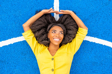 Happy young woman with Afro hairstyle lying on blue basketball court