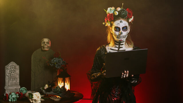 Creepy lady with halloween costume holding laptop, posing on tranditional mexican holiday in studio. Wearing skull make up body art on day of the dead, using wireless pc to browse internet.