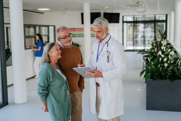 Mature doctor talking with his senior patients at hospital corridor.